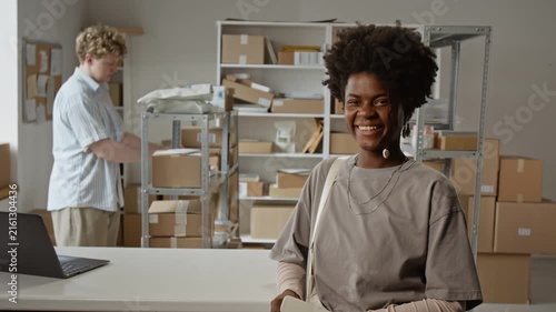 Portrait of smiling African American young adult woman standing in e-commerce warehouse while Caucasian colleague packing boxes. Footage ideal for small business and logistics marketing