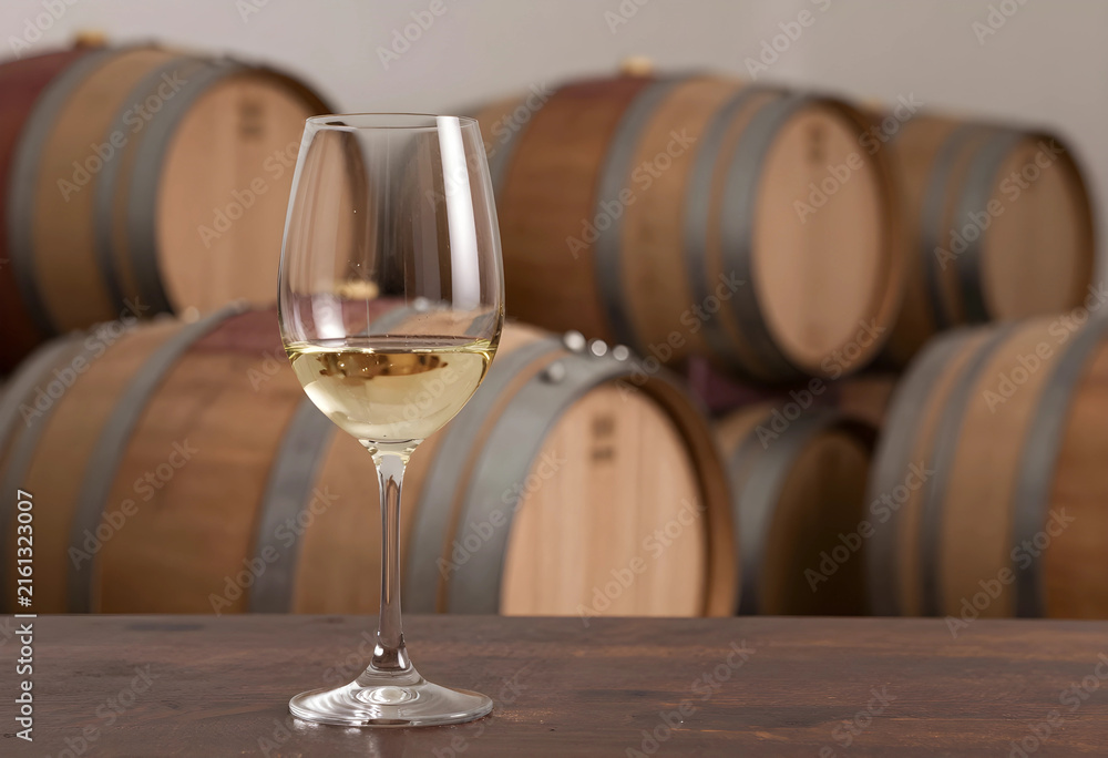 Naklejka premium Wine tasting and winemaking process, glass of white wine on a wooden table with oak barrels in a cellar