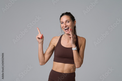 Cheerful woman in brown sportswear pointing finger at copy space on gray background