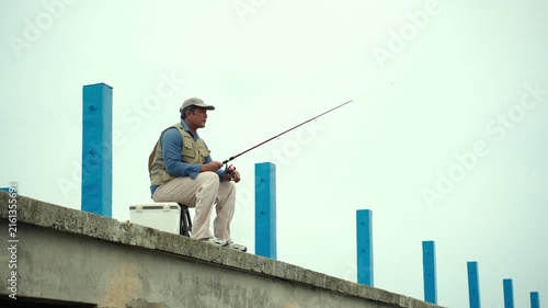 Happy senior man sitting by the sea and fishing with a rod after retirement. Active aging, healthy longevity, outdoor leisure, fulfilling retirement. Life insurance and financial security concept.
