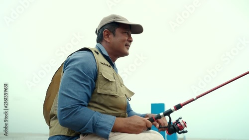 Happy senior man sitting by the sea and fishing with a rod after retirement. Active aging, healthy longevity, outdoor leisure, fulfilling retirement. Life insurance and financial security concept.
