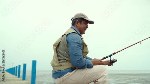 Happy senior man sitting by the sea and fishing with a rod after retirement. Active aging, healthy longevity, outdoor leisure, fulfilling retirement. Life insurance and financial security concept.
