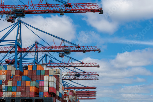 Container gantry cranes loading freight boxes on cargo ship at port terminal under blue cloudy sky, global shipping and logistics concept