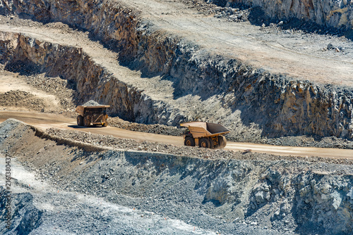 Large industrial mining trucks transport heavy copper ore along a dirt haul road cut into the rocky terraced walls of a massive open pit mine in Chile.