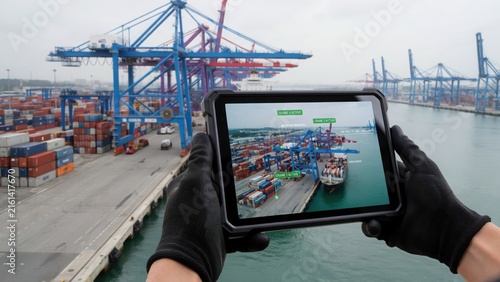 Person wearing black gloves holding a rugged tablet with augmented reality overlay showing data and information about a busy shipping port
