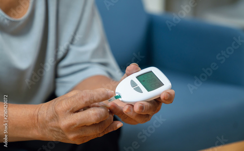 Elderly Asian woman using a blood glucose meter while sitting on a sofa at home, diabetes care and health monitoring.