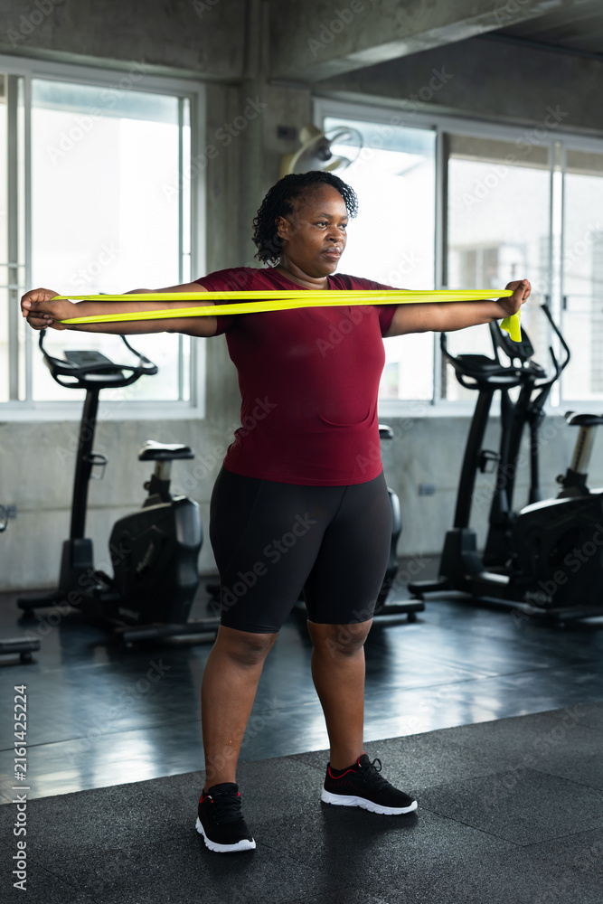 Fototapeta premium Plus size African American woman stretching arms with green resistance band in modern gym, fitness lifestyle photo for shoulder mobility, healthy workout, body confidence and wellness motivation.