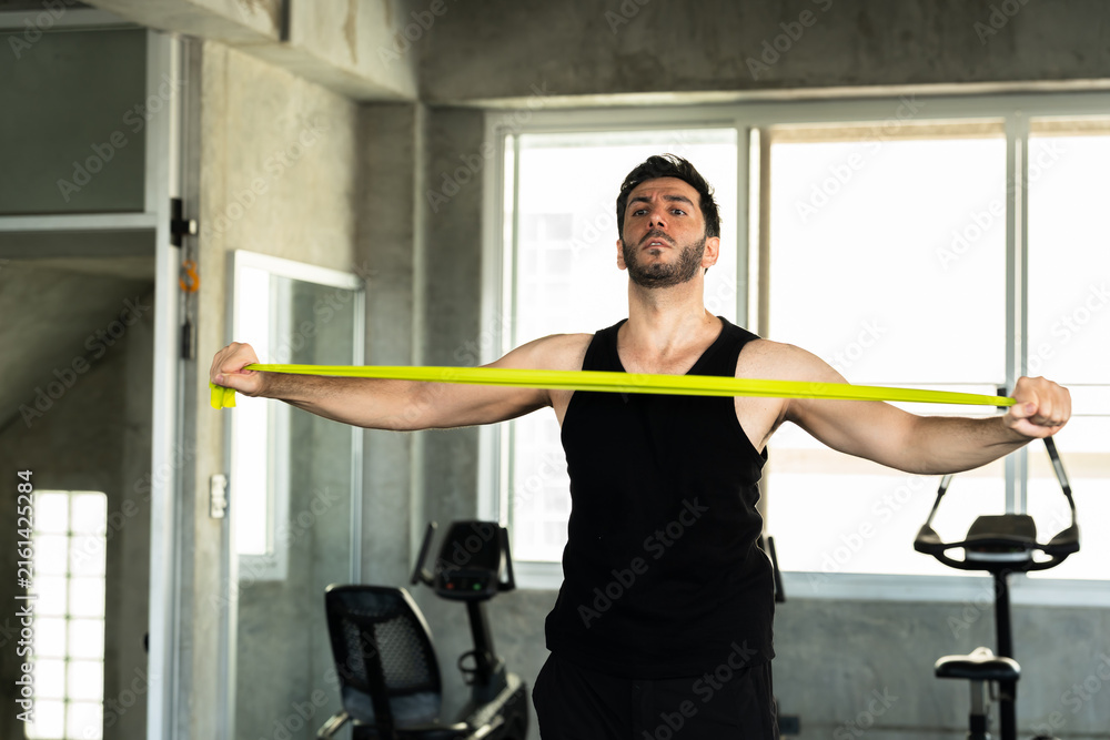Fototapeta premium Adult Caucasian man extending green resistance band across upper body in modern gym, shoulder mobility stretch before workout, active male athlete wellness routine for personal training gym ads