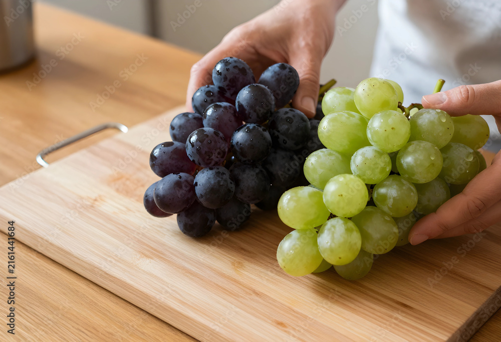Naklejka premium Healthy eating and fresh fruit concept, hands holding green and red grapes on a wooden cutting board in a kitchen