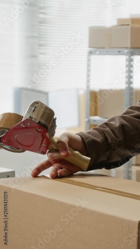 Vertical video of Caucasian young adult male sealing cardboard box with tape dispenser and sticking delivery label. Footage is ideal for e-commerce logistics and small business shipping promos