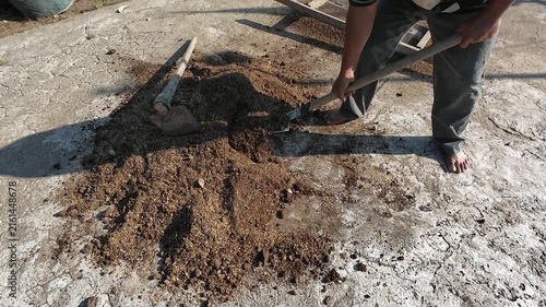 A barefoot farmer mixing organic planting media composed of soil rice husk and animal manure compost using a shovel on concrete ground preparing fertile soil for sustainable agriculture