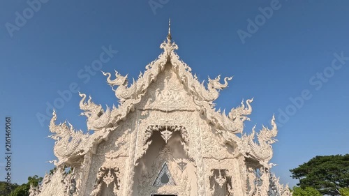 Wide shot of a white Buddhist temple in northern Thailand at midday, showcasing its handcrafted details and bright facade against a clear blue sky.