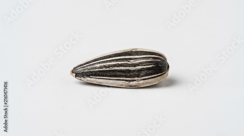 Single sunflower seed on plain white background, elongated oval shape with pointed end, striped black and beige shell, minimal composition highlighting texture and detail.