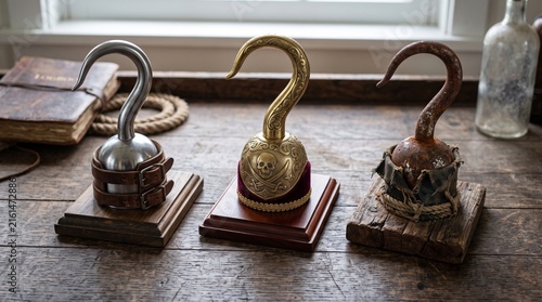 Three metal hooks on wooden surface: silver with light base, ornate gold with dark brass band, rusted worn hook, nautical background with rope, map, and brass object.