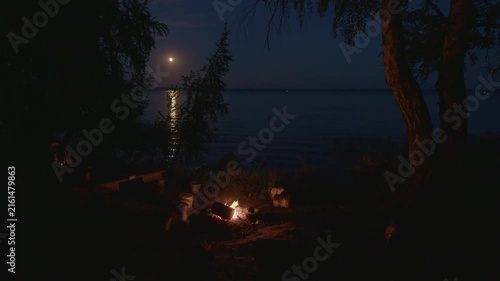 Bonfire burns on the coastal campsite surrounded by trees and with the Moon rising on the background