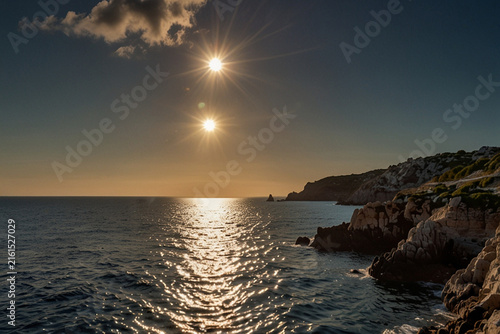Double sun phenomenon over coastal landscape with dark rocky cliffs and shimmering ocean water under a clear sky during evening golden hour.