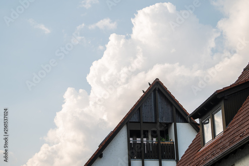 Sich auftürmende weiße Wolkengebilde vor blauem Himmel über Hausgiebel mit Rabenvögeln und Ziegeldach mit Fenster in Dachgaube in Dachgeschosswohnung in Neckartenzlingen.