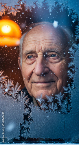Elderly man looking through frosted glass window with ice patterns and warm orange light in background during winter
