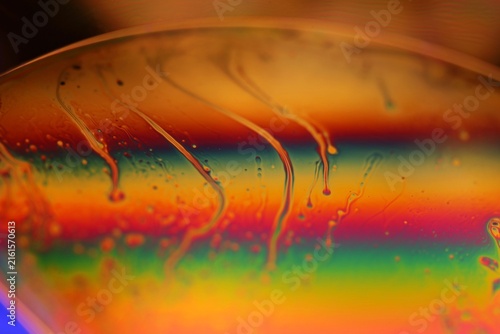 Vibrant iridescent liquid film with dripping tendrils and rainbow spectrum. A smooth, curved fluid surface showcases vivid rainbow interference bands in red, orange, green, and blue.