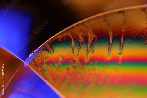Iridescent fluid film dome with dripping tendrils and vibrant rainbow interference. Intense warm amber backlighting illuminates a curved fluid dome horizon