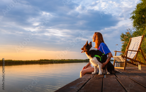 Wide shot of woman and her dog sitting on wooden pier looking at sunset over lake.