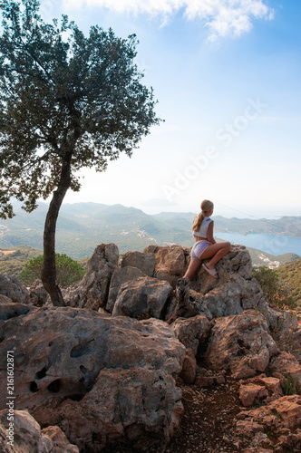 Back view of a blonde little girl sitting on rocks under a lone tree and looking at a beautiful mountain valley and sea landscape from a high viewpoint