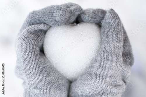 Top view close up of hands in grey knitted mittens holding a snow heart on a white snowy background