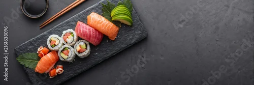 Obraz  z motywem Assorted sushi and sashimi served on a slate plate with soy sauce, chopsticks, and avocado. Japanese cuisine concept with dark copy space for menu design and food promotion.