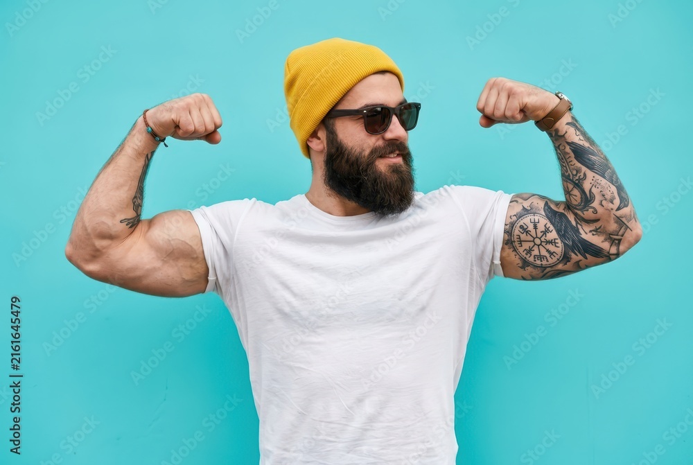 Naklejka premium Strong Muscular Bearded Man with Tattoos Flexing Biceps in White T-shirt and Yellow Beanie on Blue Background