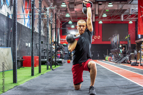 Focused young man performing a kneeling kettlebell press exercise at gym