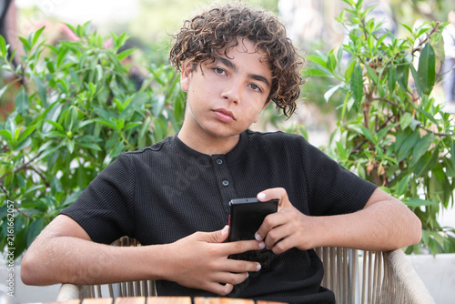 Young curly-haired boy using a smartphone outdoors, sitting at a table surrounded by greenery. Modern childhood, mobile technology, social media, digital communication and teenage lifestyle concept.