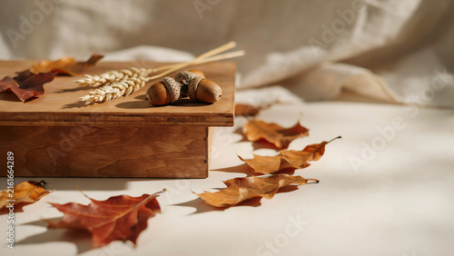 Acorns and wheat stalks arranged on a wooden surface with fallen autumn leaves scattered around during soft natural light
