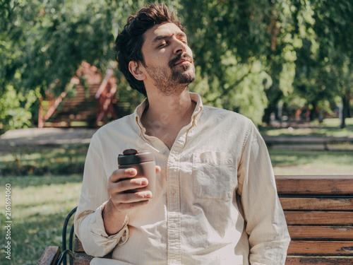 Relaxed adult man enjoying a takeaway coffee while sitting on a wooden bench in a green park while taking in sunlight on his face.