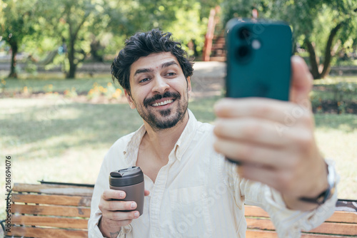 Happy adult man taking a selfie with his smartphone while sitting on a park bench outdoors. The bright lifestyle scene represents social media, mobile photography, outdoor leisure, communication and p