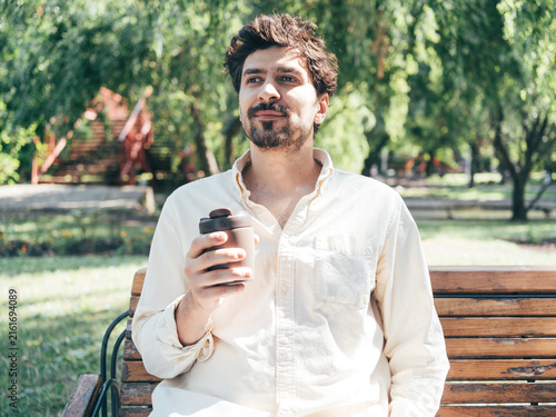 Smiling adult man enjoying coffee while talking on his smartphone in a sunny park. The lifestyle image combines outdoor relaxation, communication, technology, social connection and everyday leisure.