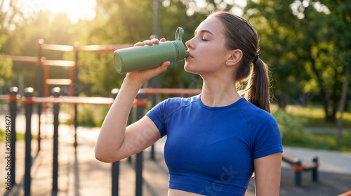 Young woman drinks water from reusable bottle during outdoor fitness training in warm sunlight