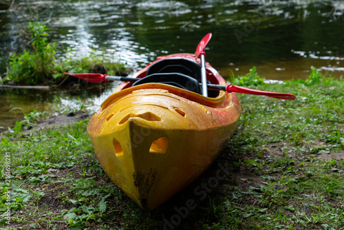 Fotografie Colorful yellow and orange kayak with paddles beside a forest stream, outdoor wa