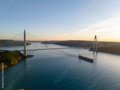 Aerial vertical view of Yavuz Sultan Selim Bridge spanning Bosphorus Strait in Istanbul, Turkey. Modern cable-stayed bridge with cargo ship navigating through sea at golden sunset.