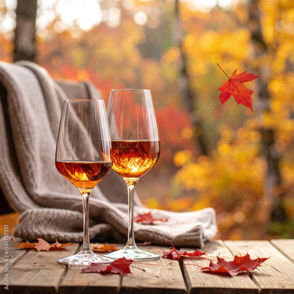 Obraz premium Two glasses of golden wine rest on a wooden rustic table surrounded by red leaves in a cozy autumn forest, evoking warmth and romantic seasonal tranquility with nobody present.