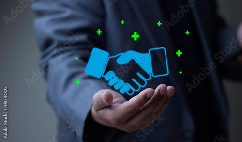 Fotografía Business partnership, corporate synergy, and strategic alliance concept with businessman hand holding glowing handshake icon surrounded by green plus symbols hologram for successful deal