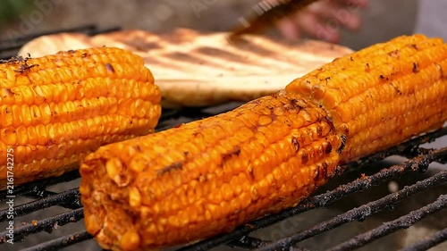Close-up of golden seasoned corn cobs being turned with metal tongs on a outdoor barbecue grill