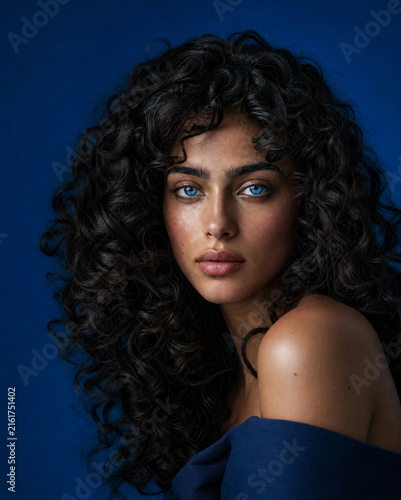 Stunning beauty editorial portrait of a young woman with curly dark hair and striking blue eyes against a monochrome background