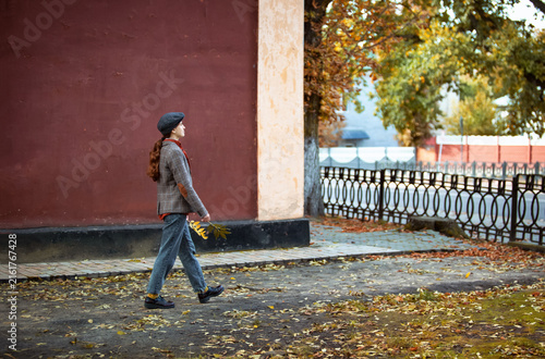 Young stylish woman wearing a flat cap and tweed jacket holding autumn leaves while walking along a city street next to a red building wall. Autumn street fashion, red brick wall, fall foliage leaves