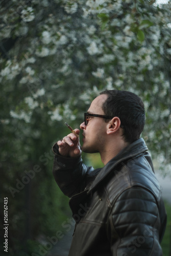 Canvas Print Side profile of a young man wearing sunglasses and leather jacket smoking a cigarette outdoors near blooming spring trees