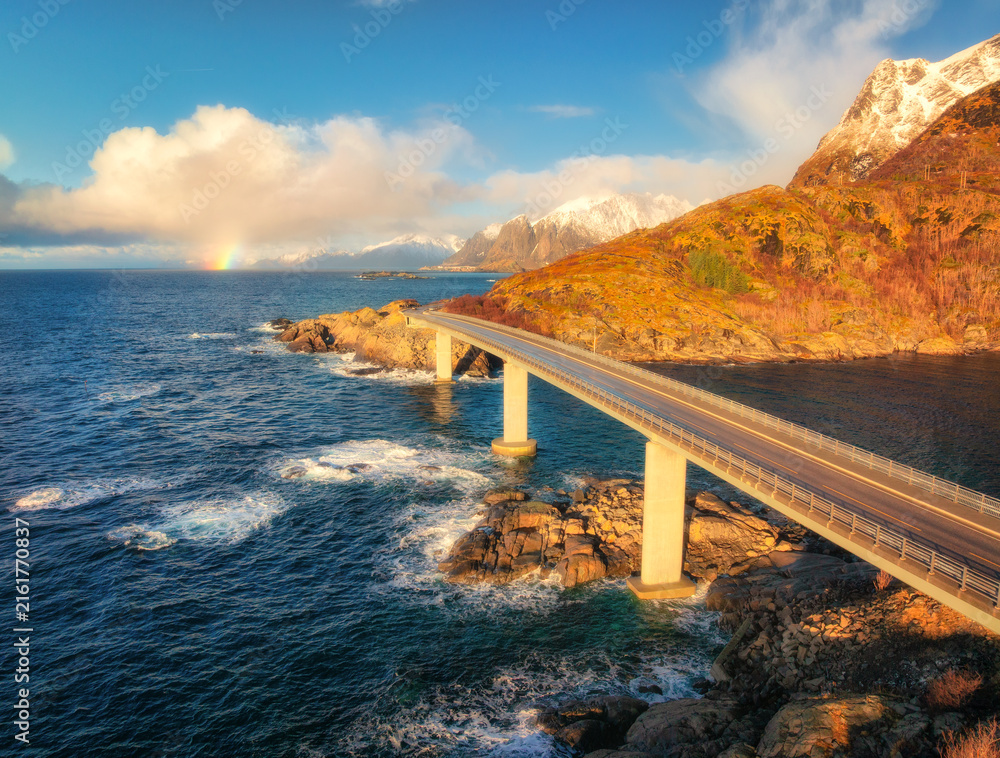 Obraz premium Aerial view of bridge, sea with waves and mountains at sunrise in Lofoten Islands, Norway. Landscape with beautiful road, water, rocks, blue sky with clouds and rainbow. Top view from drone