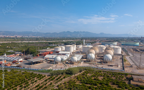 Overhead view of a large petrochemical storage facility featuring spherical LPG tanks and cylindrical oil tanks surrounded by agricultural land and mountains