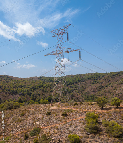 Steel electricity pylon standing on a rocky scrubland hillside with power lines stretching across a partly cloudy blue sky above pine-covered Mediterranean slopes