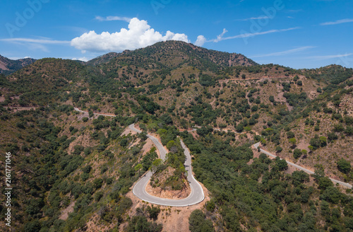 Aerial view of a serpentine mountain road with sharp hairpin curves cutting through forested hillsides, leading toward a distant summit with communication towers under a partly cloudy sky