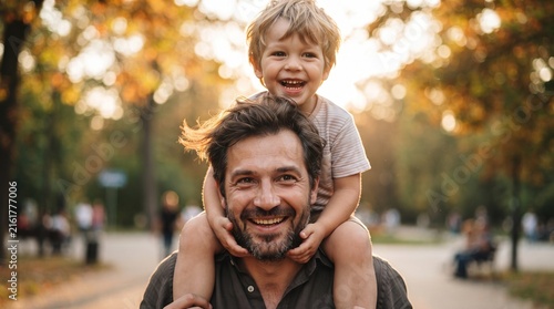 A father carries his joyful son on his shoulders, sharing a warm embrace and a radiant smile amidst the golden hues of an autumn park.