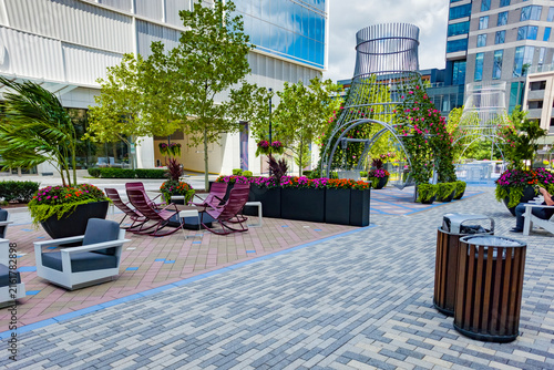 Wide paved public plaza connects modern shops, residences, seating, and flower displays.